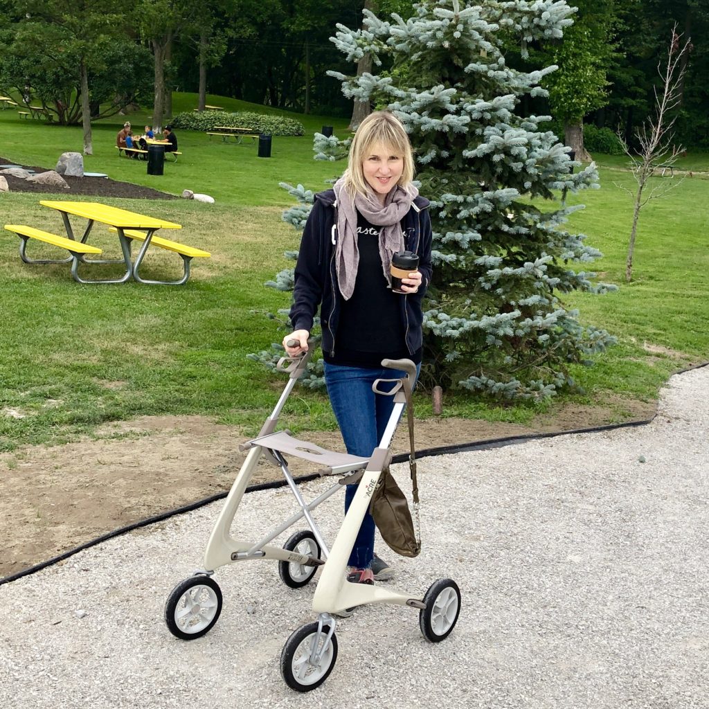 Ardra, a babe with a mobility aid, stands behind her oyster coloured rollator from byAcre. She is in a park, looking chill, holding a coffee, with one leg crossed casually over the other.