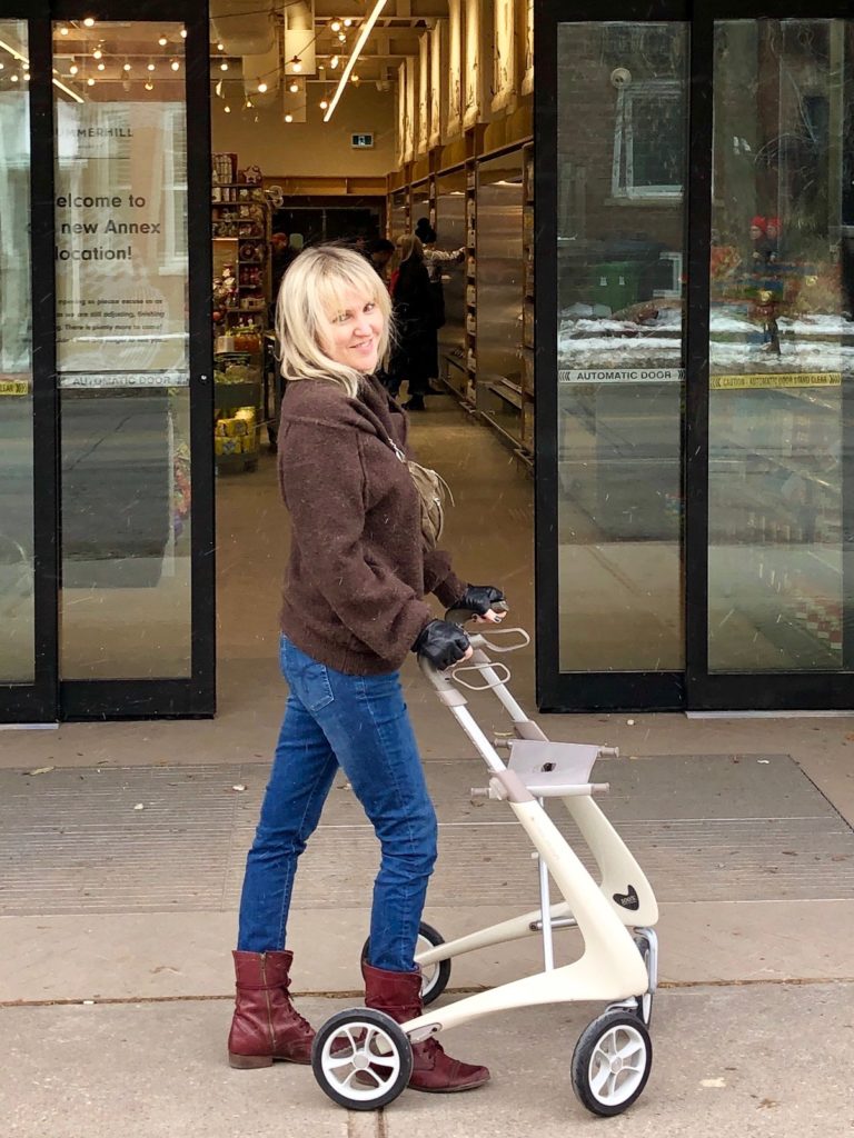 Ardra stands with her byAcre rollator. The oyster white rollator is sleek and stylish, just like Ardra who's wearing skinny jeans tucked into wine colored red leather boots and a chunky brown turtle-neck.
