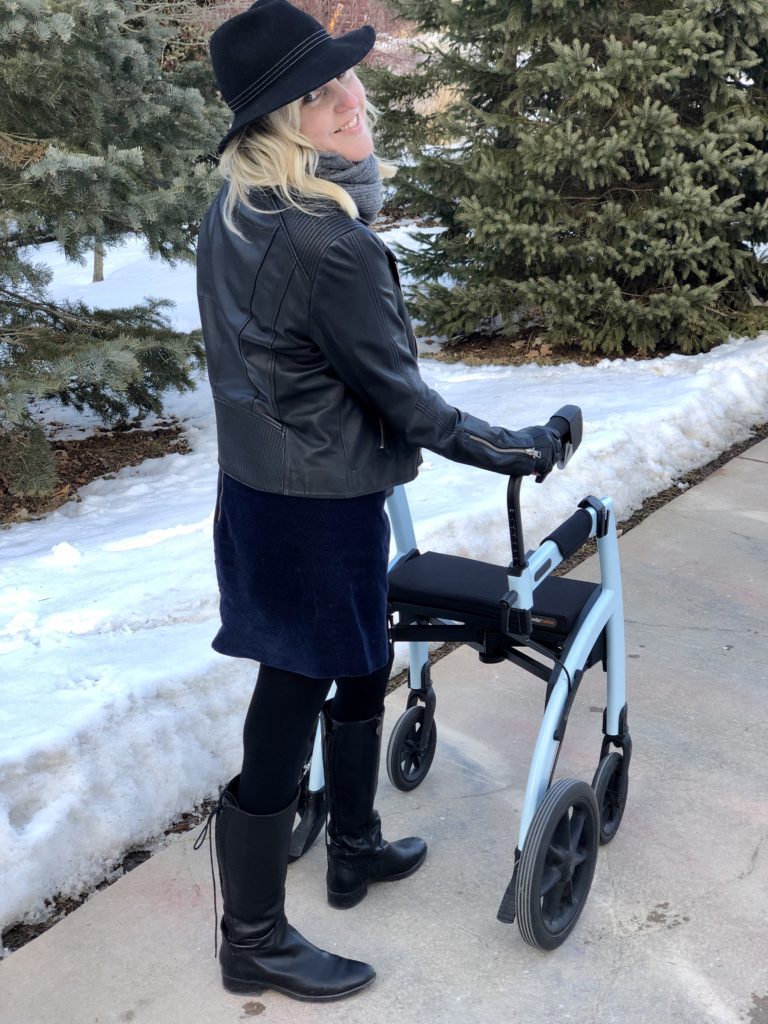Ardra, a white woman with blonde hair, stands behind her Rollz in rollator mode. She's looking back over her shoulder and smiling. She looks cool in her leather jacket and her confidence with the Rollz Motion only makes her look cooler.