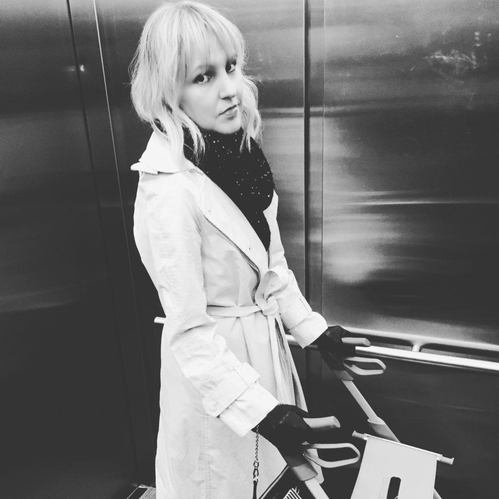 A white woman with MS is in an elevator. She is wearing a trench coat and looks irritated.